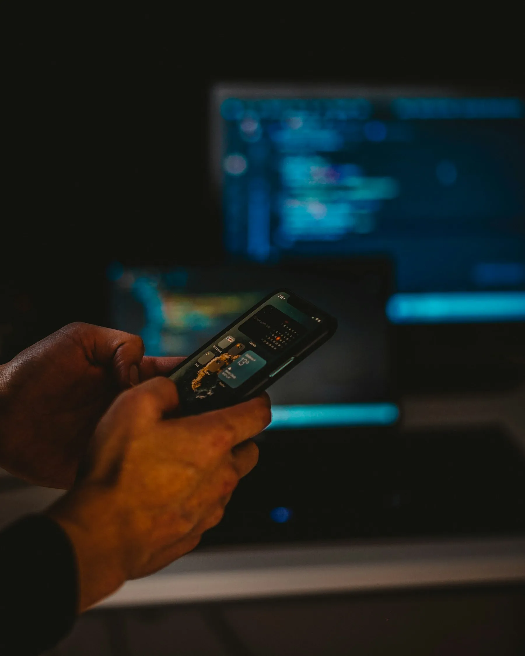 Mãos segurando um celular diante de telas com código, em um ambiente escuro